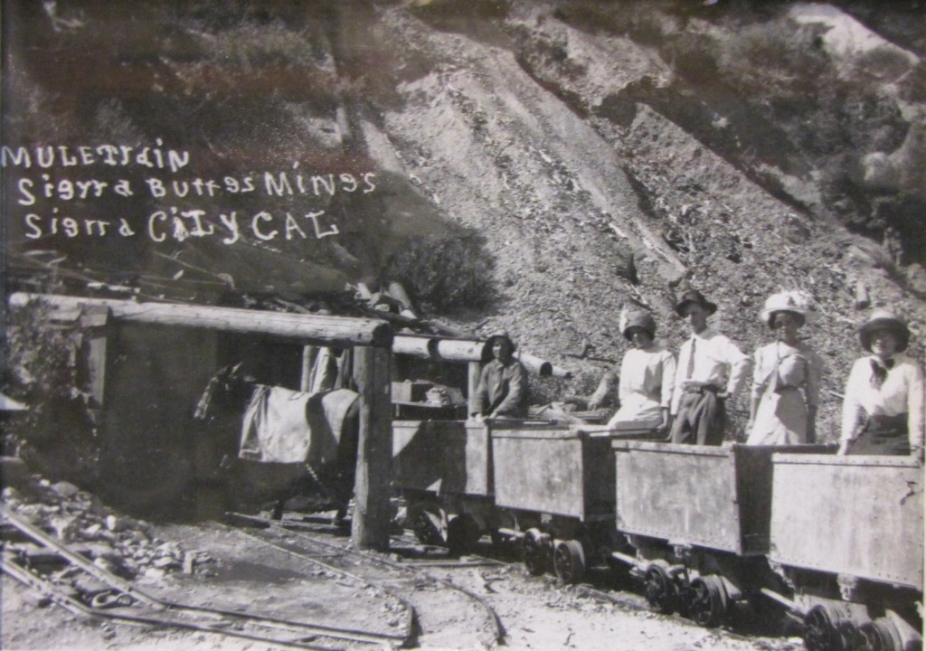 Ladies in large hats riding ore cars into the mine.